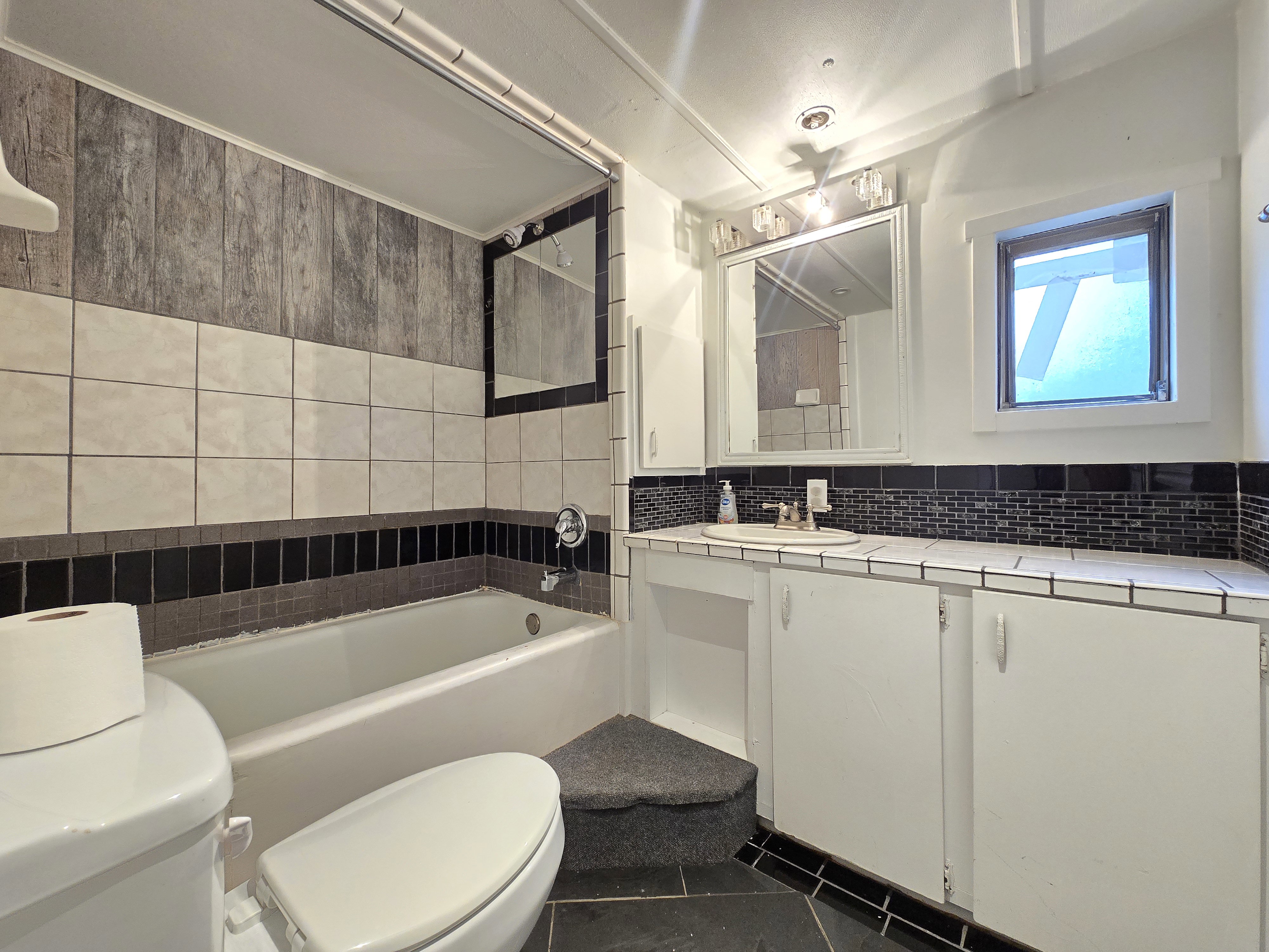 Bathroom featuring a built-in tub with a tiled surround, a vanity with an integrated sink and white cabinetry, a large wall-mounted mirror, and black tile flooring