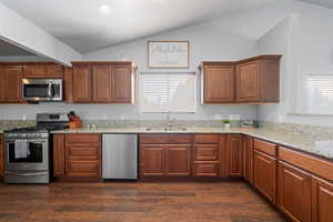 Kitchen with stainless steel appliances, wood finish cabinets, light stone counters, dark wood-style flooring, and vaulted ceiling