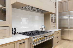 Kitchen featuring light wood finish cabinetry, high quality appliances, light stone counters, decorative backsplash, and glass insert cabinets