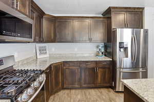 Kitchen featuring stainless steel appliances, dark wood finish cabinetry, light stone countertops, a textured ceiling, and light wood-style floors