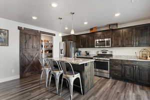 Kitchen with a barn door, dark wood finish cabinets, stainless steel appliances, light stone countertops, and an island with sink