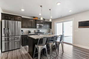 Kitchen featuring stainless steel appliances, light stone countertops, an island with sink, a breakfast bar, and dark wood-style flooring