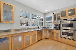 Kitchen with glass insert cabinets, stainless steel appliances, light wood-style flooring, light stone counters, and recessed lighting