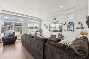 Living room with wood finished floors, a glass covered fireplace, a raised ceiling, and recessed lighting