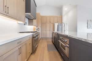 Kitchen featuring stainless steel appliances, light stone countertops, light wood-type flooring, two tone cabinets, and lofted ceiling