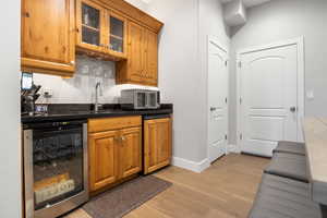 Indoor wet bar with beverage cooler, glass fronted cabinets, light wood-type flooring, wood finish cabinets, and dark stone countertops