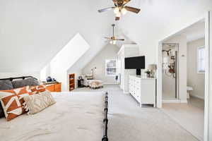 Bedroom with vaulted ceiling, multiple windows, light carpet, connected bathroom, and ceiling fan