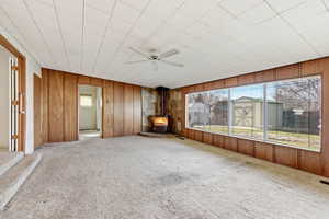 Unfurnished room with a ceiling fan, a wood stove, carpet floors, and wooden walls