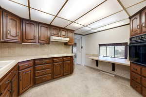 Kitchen featuring black oven, light countertops, light carpet, wallpapered walls, and dark wood finish cabinetry