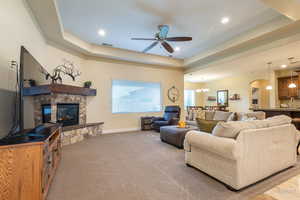 Living room featuring a ceiling fan, arched walkways, a raised ceiling, light colored carpet, and a stone fireplace