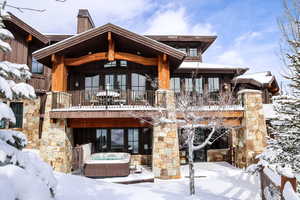 Snow covered house featuring a balcony, a hot tub, a chimney, a patio area, and stone siding