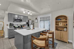 Kitchen featuring gray cabinetry, vaulted ceiling, stainless steel appliances, an island with sink, and light stone countertops