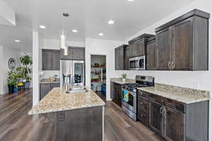 Kitchen featuring stainless steel appliances, dark wood finish cabinets, light stone counters, pendant lighting, and an island with sink