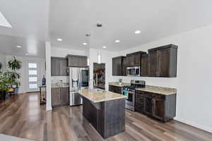 Kitchen featuring dark wood finish cabinets, stainless steel appliances, light stone countertops, a kitchen island with sink, and recessed lighting