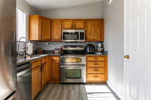 Kitchen with stainless steel appliances, wood finish cabinetry, vaulted ceiling, backsplash, and light wood-type flooring