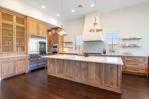 Kitchen featuring open shelves, decorative light fixtures, glass fronted cabinets, stainless steel appliances, and a kitchen island with sink