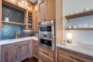 Kitchen with stainless steel double oven, light stone counters, glass insert cabinets, decorative backsplash, and open shelves
