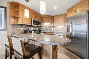 Kitchen featuring a peninsula, stainless steel appliances, light stone counters, decorative light fixtures, and light wood finish cabinets