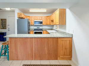 Kitchen with stainless steel appliances, light stone counters, a peninsula, light tile patterned floors, and light wood finish cabinetry