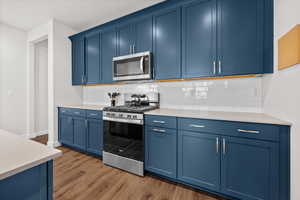 Kitchen with stainless steel appliances, blue cabinets, dark wood-style floors, backsplash, and light stone countertops