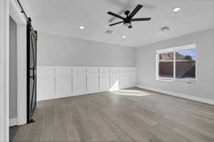 Spare room featuring a barn door, light wood finished floors, a decorative wall, ceiling fan, and wainscoting