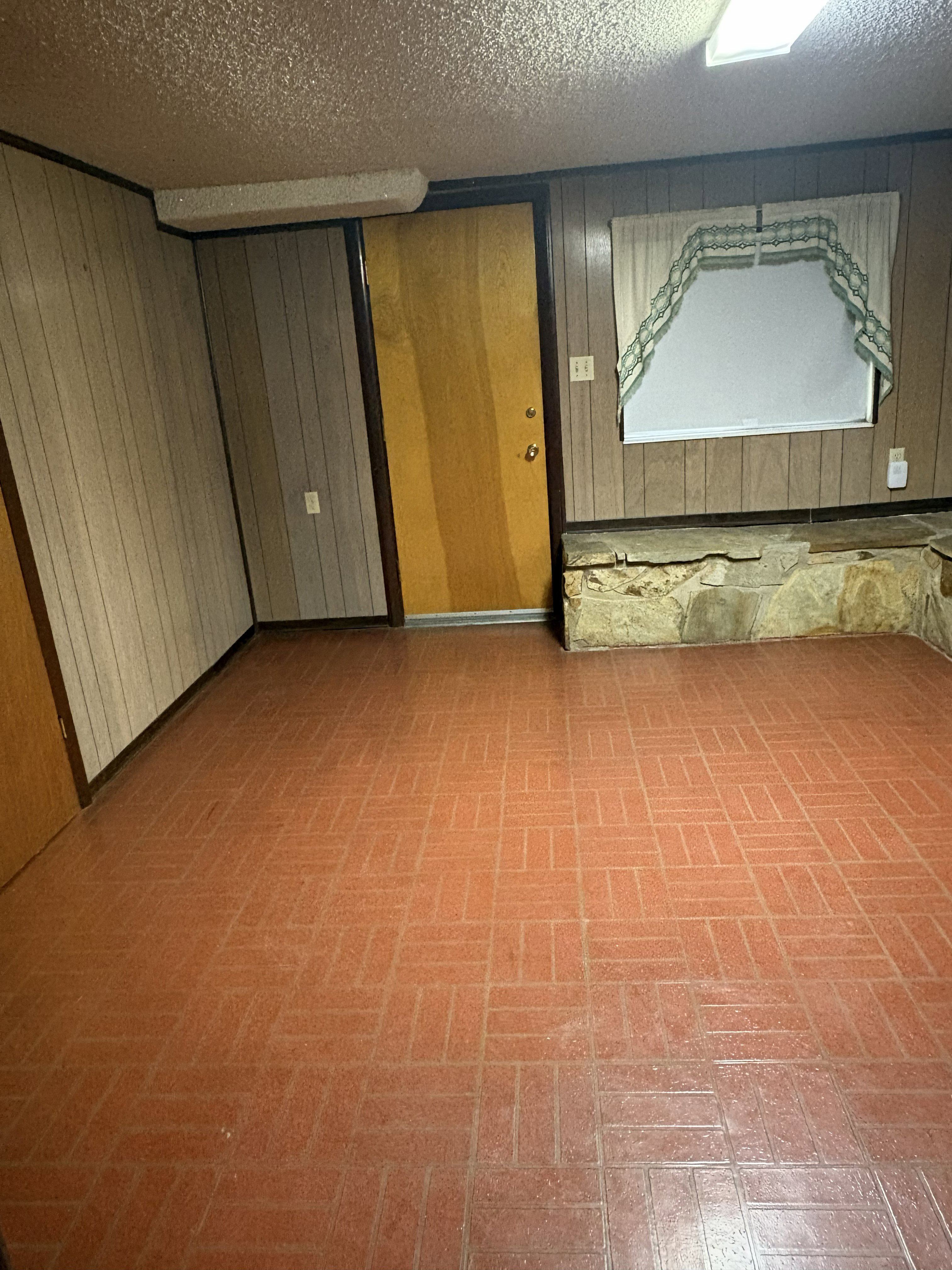 Room featuring red-toned geometric tile flooring, wood paneling, a light wood door, and a window with a stone base