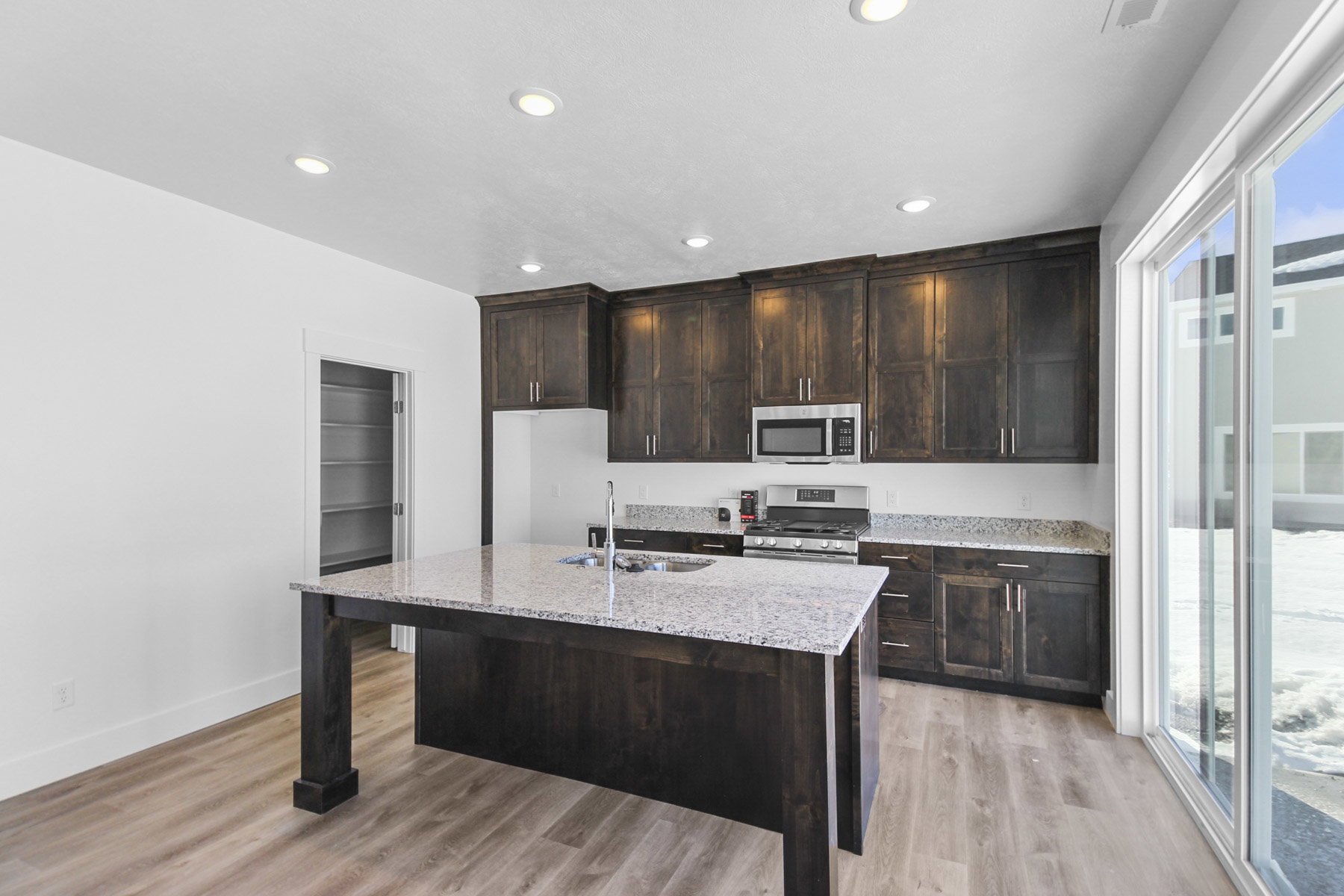 Kitchen featuring dark wood finish cabinetry, stainless steel appliances, light stone countertops, light wood-style flooring, and a kitchen island with sink
