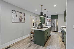 Kitchen with green cabinets, an island with sink, stainless steel appliances, light stone counters, and light wood finished floors