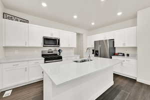 Kitchen featuring stainless steel appliances, white cabinetry, a kitchen island with sink, dark wood-type flooring, and light stone countertops