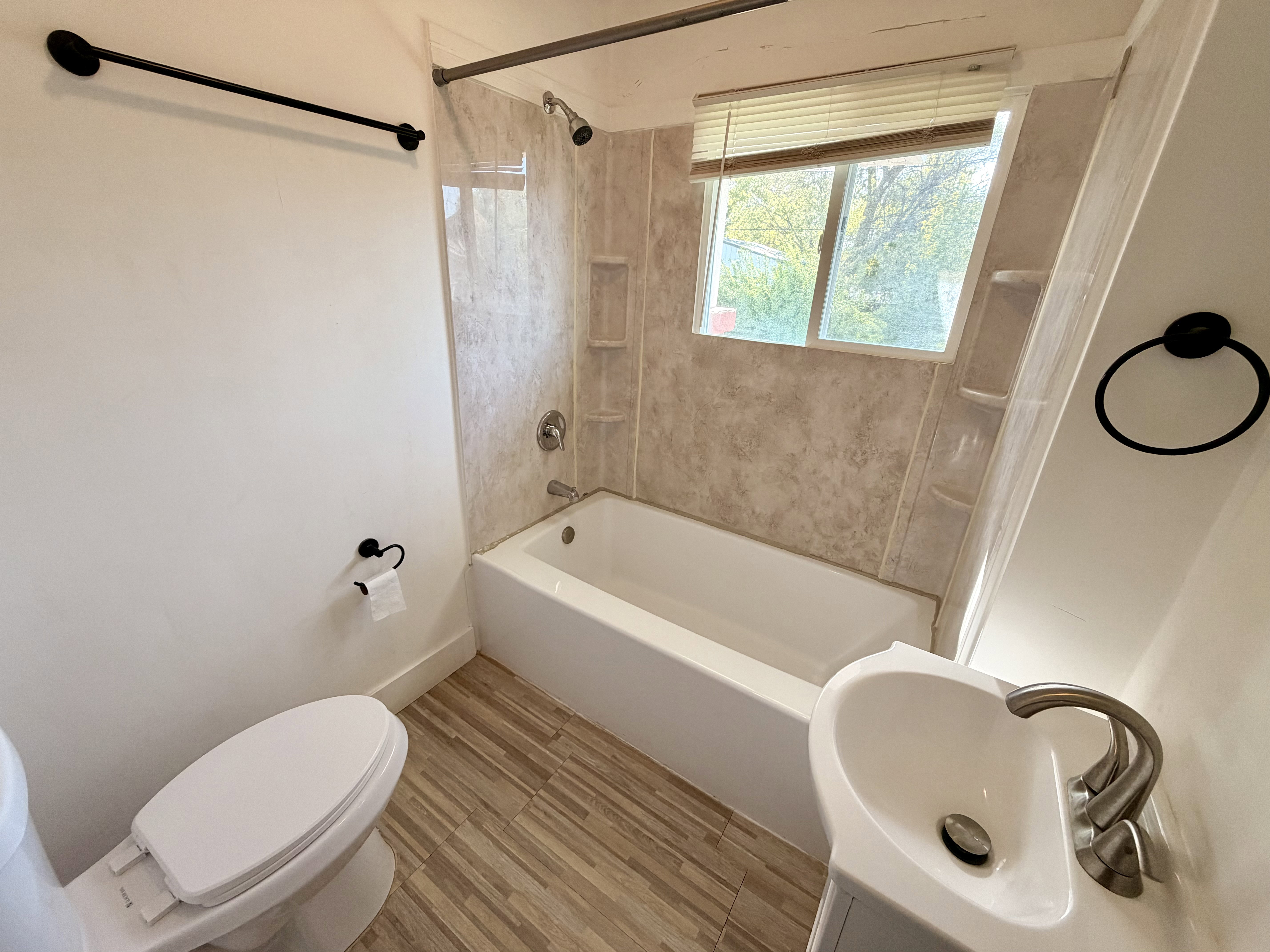 Bathroom featuring a full tub and shower combination with light-toned wall surround, a window with blinds, wood-finish flooring, and a pedestal sink with a brushed nickel faucet