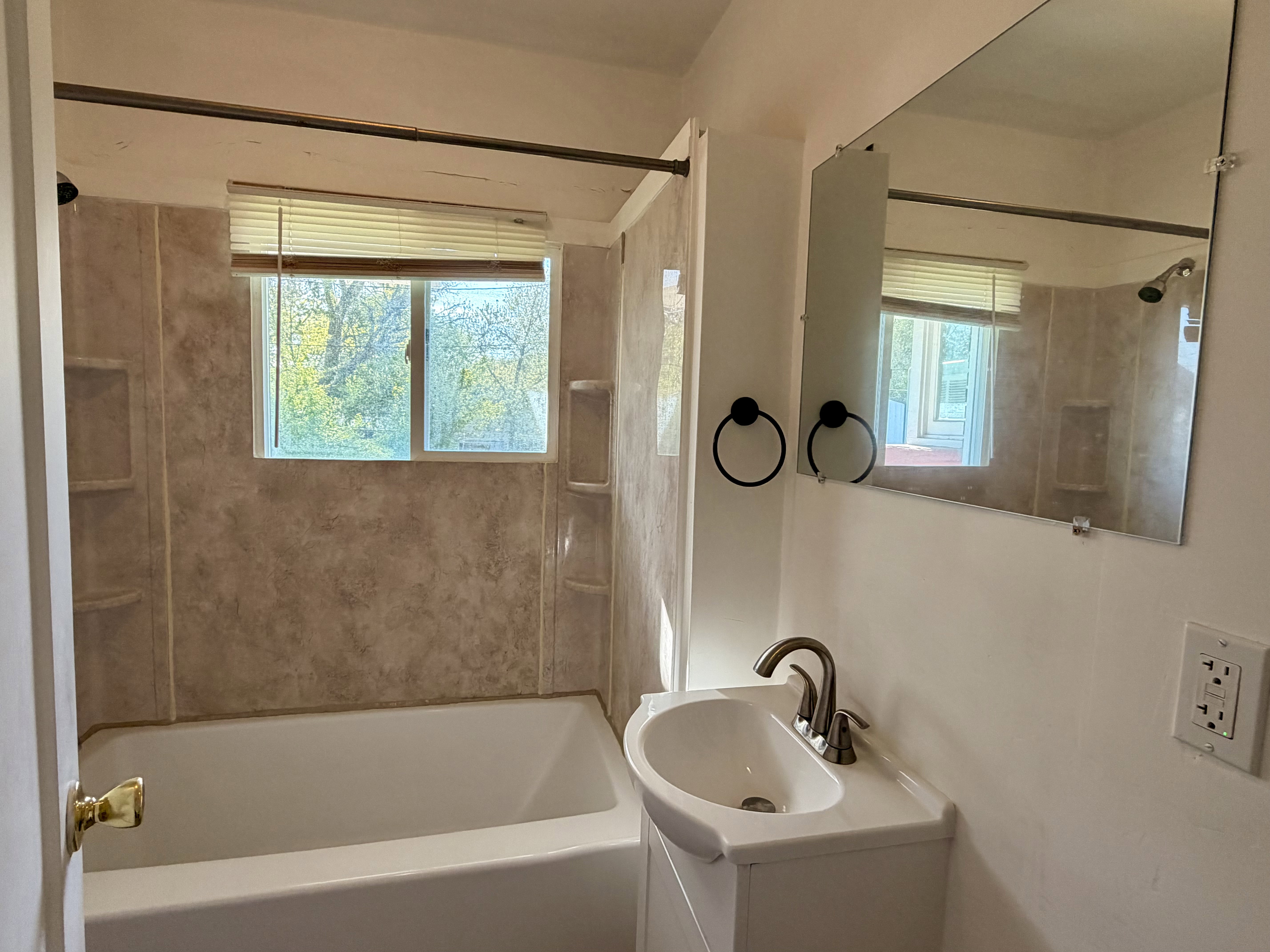 Bathroom featuring a built-in tub with textured surround, window with blinds, wall-mounted mirror, and a vanity with an integrated sink and brushed nickel faucet