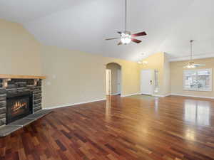 Unfurnished living room featuring a ceiling fan, arched walkways, vaulted ceiling, a stone fireplace, and dark wood finished floors