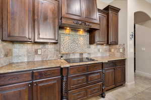 Kitchen with light stone countertops, tasteful backsplash, light tile patterned floors, black electric stovetop, and dark wood finish cabinetry