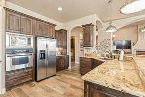 Kitchen with stainless steel appliances, dark wood finish cabinets, light stone countertops, decorative light fixtures, and light wood-style flooring