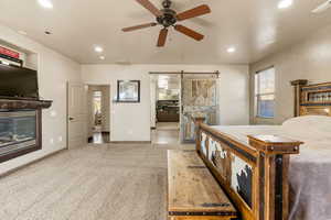 Bedroom featuring a barn door, a glass covered fireplace, ensuite bathroom, light colored carpet, and a ceiling fan