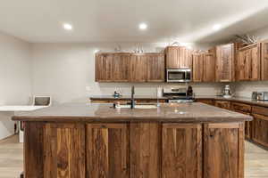 Kitchen with light wood-type flooring, a kitchen island with sink, stainless steel appliances, wood finish cabinetry, and dark stone countertops