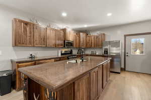 Kitchen featuring stainless steel appliances, light wood-style flooring, a center island with sink, wood finish cabinets, and dark stone countertops
