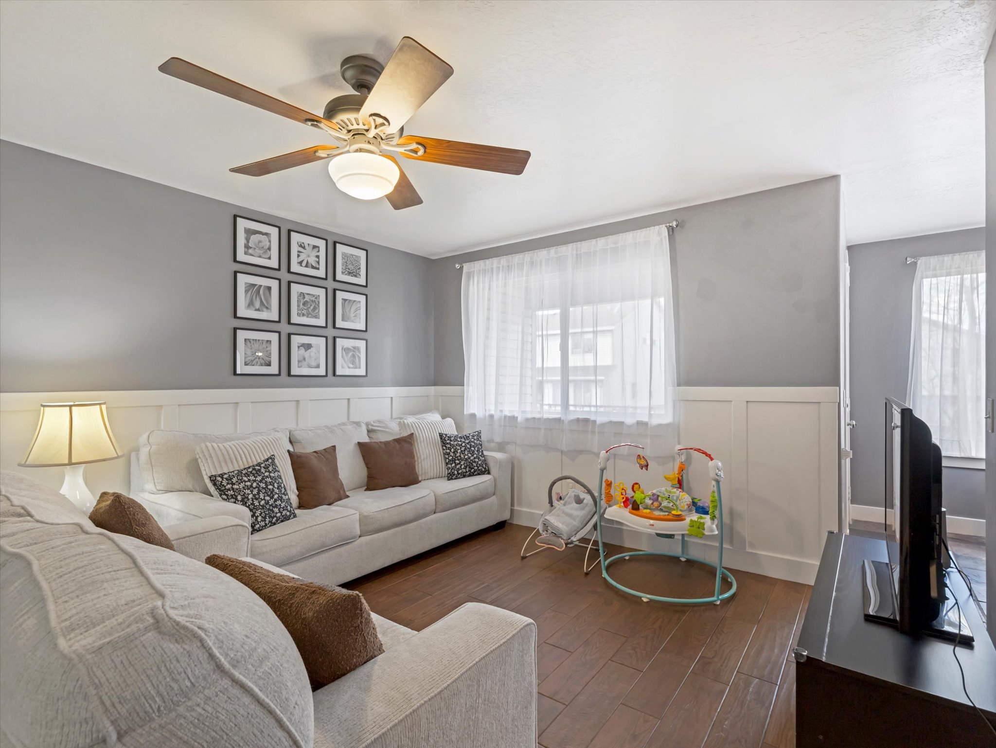 Living room with a wainscoted wall, dark wood-style flooring, ceiling fan, healthy amount of natural light, and a decorative wall