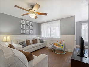 Living room with a wainscoted wall, dark wood-style flooring, ceiling fan, healthy amount of natural light, and a decorative wall