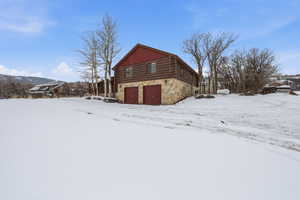 View of snowy exterior with stone siding, log exterior, and a garage