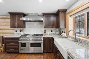 Kitchen with range with two ovens, dark wood finish cabinets, light stone countertops, and log walls