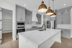 Kitchen with gray cabinets, light stone counters, stainless steel appliances, light wood-style flooring, and an island with sink