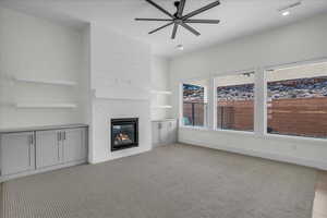 Unfurnished living room featuring a brick fireplace, a ceiling fan, built in shelves, and light colored carpet