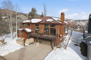 Snow covered property with a deck, a garage, driveway, a chimney, and stone siding