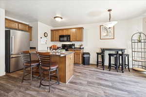 Kitchen featuring black appliances, wood finish cabinets, a center island with sink, hanging light fixtures, and dark countertops