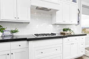 Kitchen featuring white cabinetry, glass insert cabinets, stainless steel gas stovetop, backsplash, and dark stone countertops