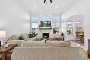 Living room with arched walkways, light wood finished floors, a tiled fireplace, ceiling fan, and a high ceiling