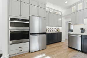 Kitchen with two tone cabinets, stainless steel appliances, light wood-style flooring, glass fronted cabinets, and light stone countertops