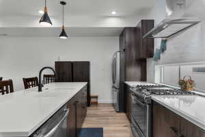 Kitchen with stainless steel appliances, dark wood finish cabinetry, pendant lighting, light wood-style floors, and light stone countertops