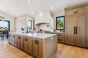 Kitchen with brown cabinetry, a center island with sink, recessed lighting, light wood-type flooring, and light stone countertops